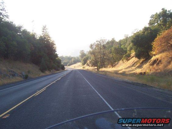 View from a vehicle driving on a two-lane road through a wooded, hilly area