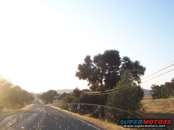 View from a vehicle driving on a rural road with trees and hills under a bright sky