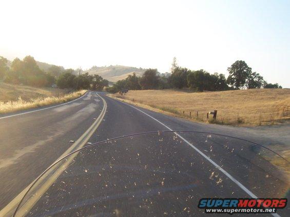 View from a vehicle looking down a rural road with dry grass and trees under a hazy sky