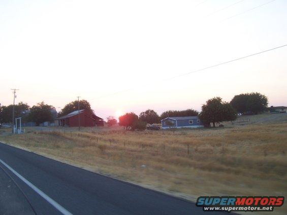 Rural landscape with a red barn, a blue mobile home, and dry grass under a sunset sky