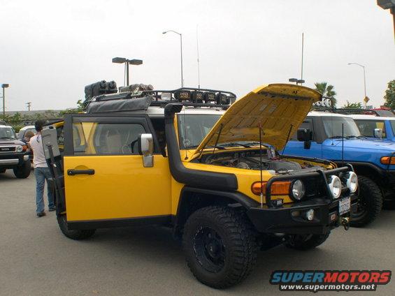 Yellow 2007 Toyota FJ Cruiser with hood open, snorkel, roof rack, and aftermarket bumper. Blue FJ Cruiser parked behind