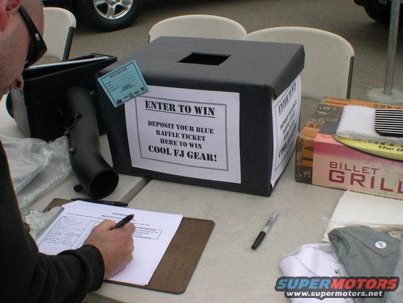Raffle box with 'ENTER TO WIN COOL FJ GEAR!' sign, clipboard with pen, and other items on a table