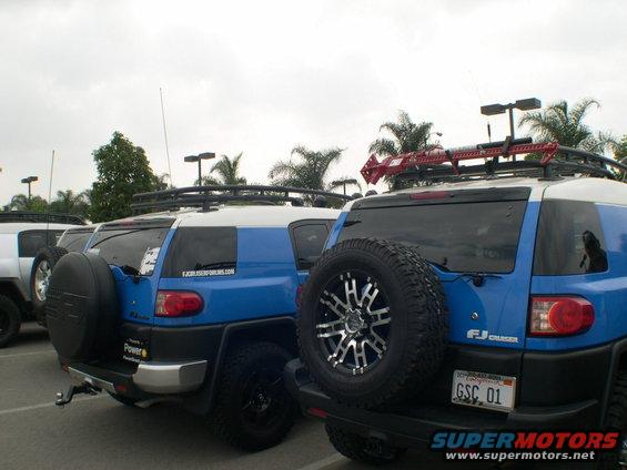 Two blue and white Toyota FJ Cruisers parked in a lot. One has a red Hi-Lift jack on its roof rack