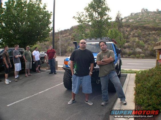 Two guys stand in front of a blue 2007 Toyota FJ Cruiser. Other people and vehicles are in the background