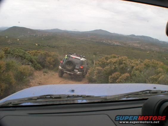 Driving behind a 2007 Toyota FJ Cruiser on a dirt road with hills and brush