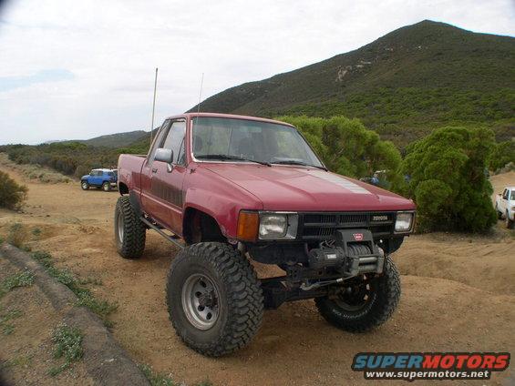 Red Toyota pickup truck modified for off-roading, parked on a dirt road with another blue SUV in the background
