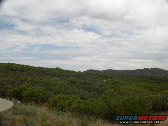 Hilly terrain covered in green bushes under a cloudy sky. A concrete structure is visible on the left