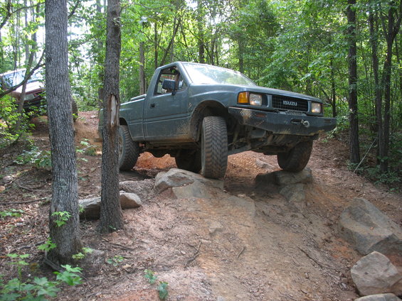 Green Isuzu pickup truck climbing over rocks on a dirt trail in the woods. Another vehicle is visible behind it