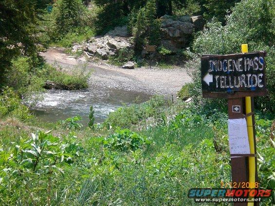 Sign pointing to Imogene Pass and Telluride next to a river in a mountainous area