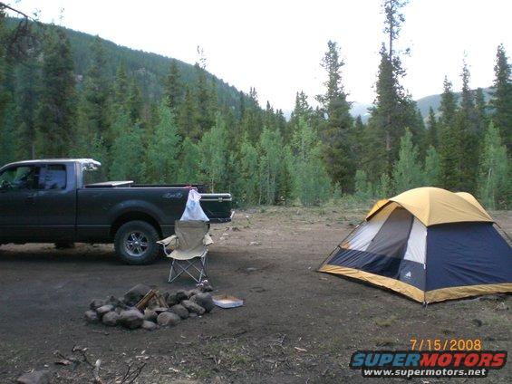 Dark gray Ford F-150 parked next to a campsite with a tent and fire pit in a wooded area