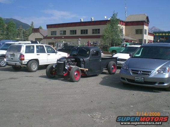 Black hot rod pickup truck with exposed engine and red wheels, parked in a lot