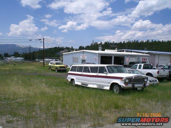 White limousine conversion with maroon stripe, parked in grassy lot next to a yellow SUV and white pickup truck