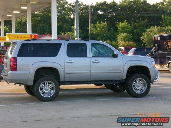 Silver SUV, looks like a Suburban, with big tires and lifted suspension. Parked at a gas station