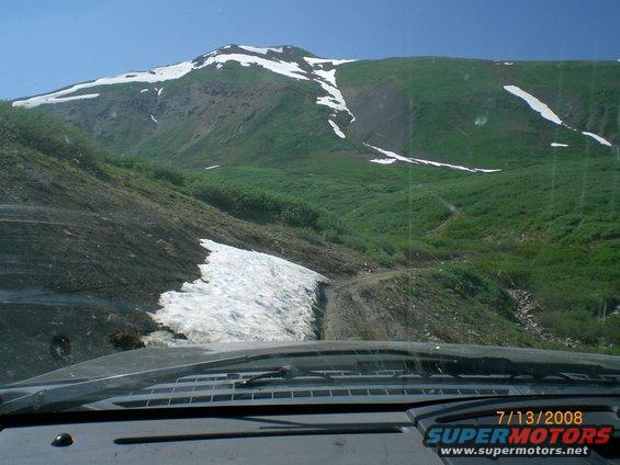 Dashboard view from a vehicle driving on a dirt road with snow patches, leading up a grassy mountain under a clear blue sky