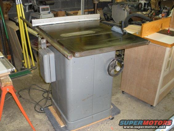 Gray table saw with metal top and a hand crank on the side, in a workshop setting