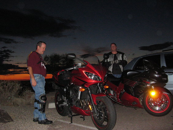 Two guys stand with their motorcycles, a red sportbike and a black cruiser, as the sun sets