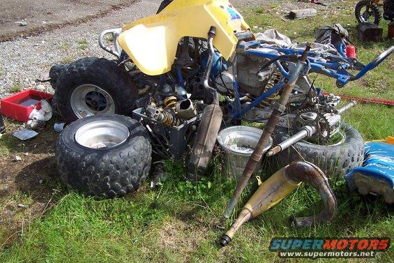 Disassembled yellow ATV with blue frame, missing body panels and engine components. Tires and exhaust pipe are visible