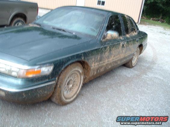 Mud-covered dark green 1995 Mercury Grand Marquis parked on gravel, with a pickup truck in the background