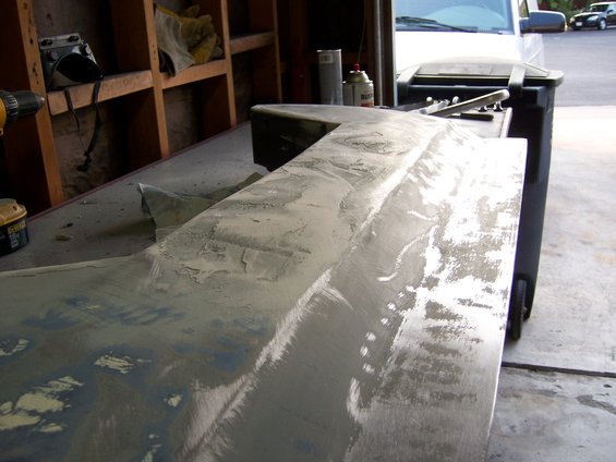 Car body panel with primer and sanding marks on a workbench, with tools and a trash can nearby