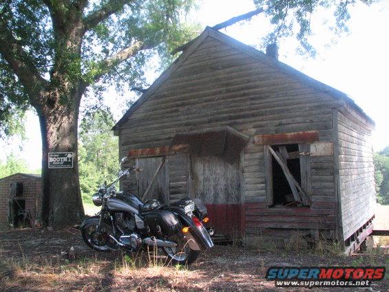 Black motorcycle parked by a rustic wooden shed. Sign on tree reads 'BOOTH 1'