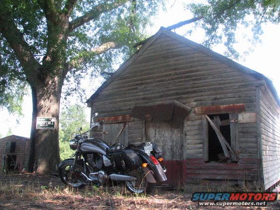 Motorcycle parked beside a weathered wooden shed under a large tree. A sign on the tree reads 'BOOTH 1'
