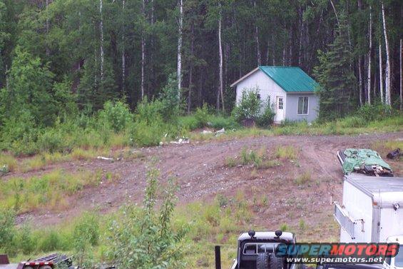 Wooded area with a small cabin, dirt road, and parts of trucks and a trailer
