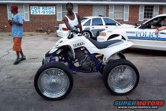 White Yamaha ATV with custom chrome wire wheels parked on pavement, with two men and a police car in the background