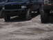 1989 Ford Bronco - Two lifted trucks, a black pickup and a red SUV, are parked on sandy ground.