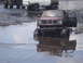 1989 Ford Bronco - A muddy GMC pickup truck sits in water with its door open, while other trucks are visible in the background.