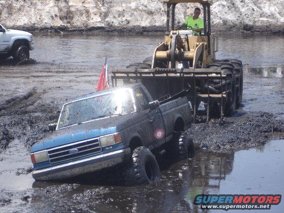 Blue Ford pickup truck stuck in mud, being pulled by a yellow front-end loader. A red and white flag is visible
