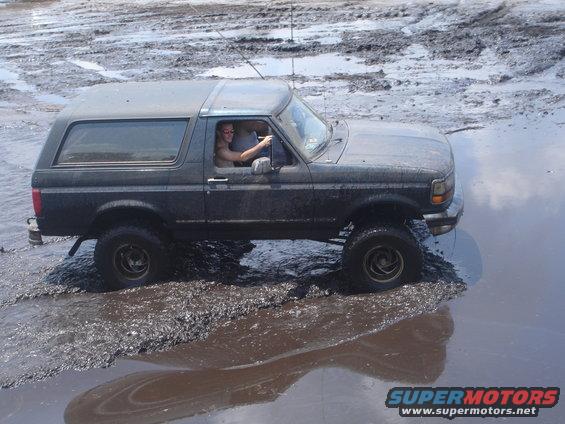 Dark gray 1989 Ford Bronco driving through mud, driver visible