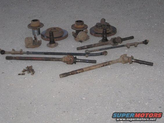Assorted rusty vehicle front axle and steering components laid out on a concrete surface