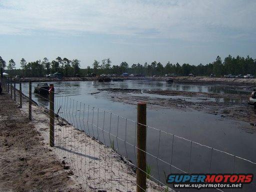 Muddy water scene with several vehicles partially submerged, trees in background, fence in foreground