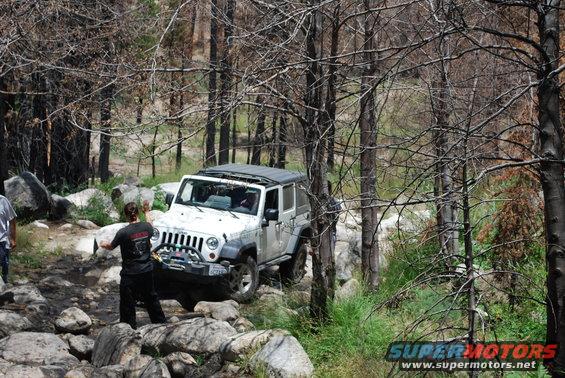White Jeep Wrangler navigating a rocky trail in a wooded area. A person is standing near the front of the Jeep