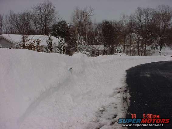 Snowy landscape with a road and a large snowbank, with houses and trees in the background