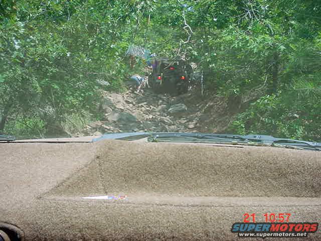 Off-road vehicle climbing a steep, rocky hill, surrounded by trees. View from inside another vehicle