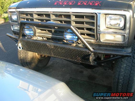 Front end of a black Ford truck with "MUDD DUCK" painted on the hood, custom bumper, and blue auxiliary lights. Grille suggests 1980s model