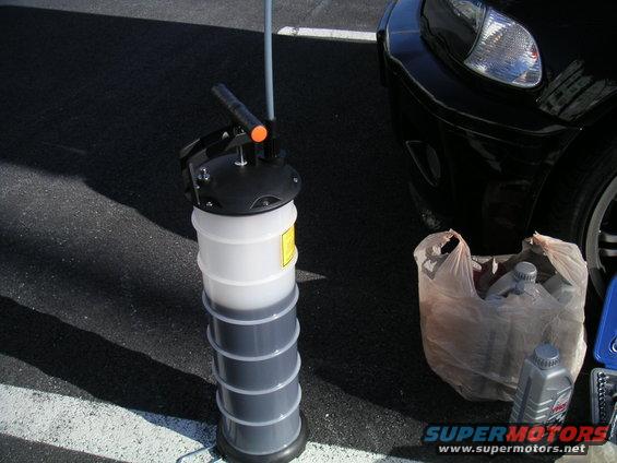 Oil drain pan filled with dark fluid, next to a plastic bag with oil bottles and a car's front fender