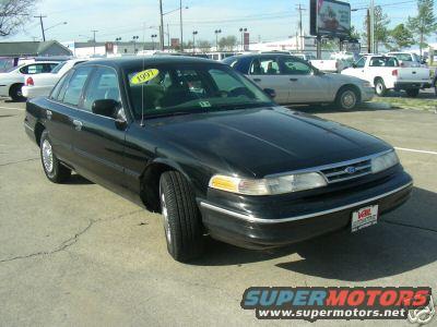 Black Ford Crown Victoria parked outside with a yellow 1997 tag on the windshield. Other cars and trucks are in the background