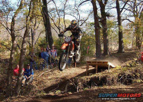 Orange dirt bike airborne over a dirt track, rider in full gear