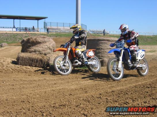 Two dirt bike riders on a dirt track with hay bales and bleachers in the background