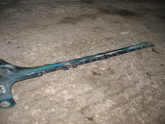 Close-up of a dark teal, metal vehicle frame component with some rust and wear, lying on a concrete surface
