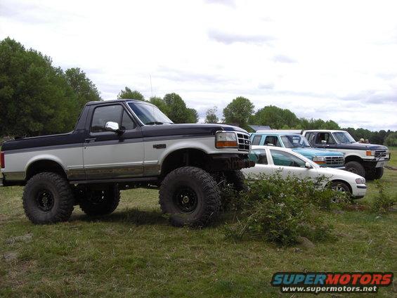 Lifted two-tone pickup truck with large tires, 3/4 front view. Other vehicles in background