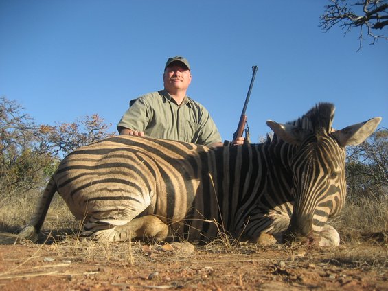 Hunter poses with a zebra he shot. He is wearing a cap and holding a rifle