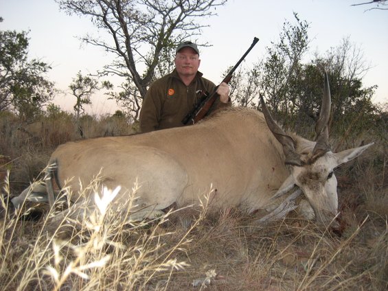 Man in hunting attire holding a rifle over a large dead antelope lying on dry grass and brush