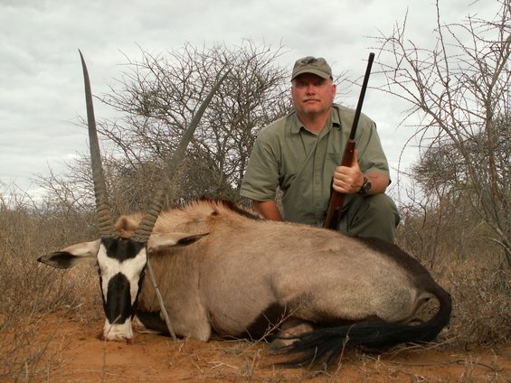 Hunter kneels behind a fallen oryx antelope, holding a rifle. Dry brush and trees in background