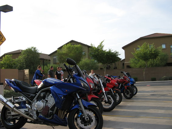 Row of parked motorcycles, blue Yamaha in foreground, red and blue motorcycles behind, people standing near them, suburban houses in background