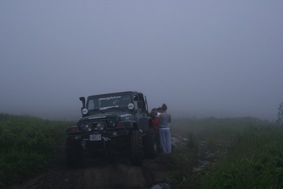 Jeep Wrangler with snorkel and off-road tires parked on a muddy track in fog. People stand near the passenger side