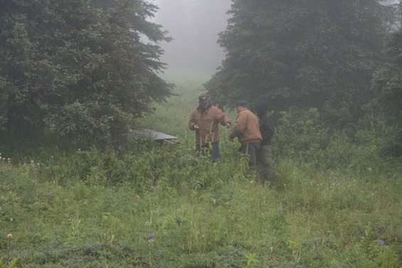 Three men stand in a foggy field near a partially visible metal structure