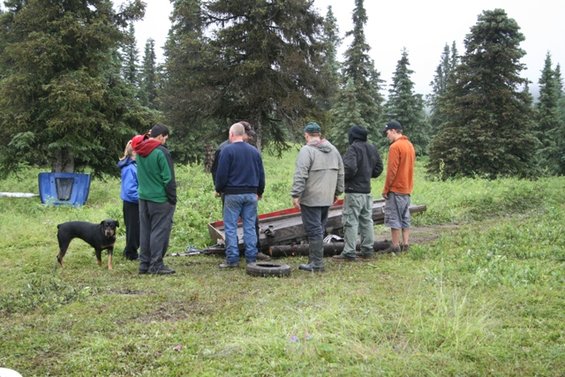 Group of people and a dog gathered around a wrecked vehicle in a wooded area